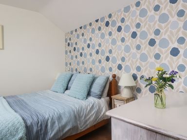 A bedroom with a bed with blue pillows and striped bedding a nightstand with a lamp and a table with a vase of flowers at The Wagon House in Polyphant near Launceston