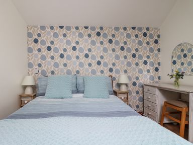 A bedroom with a bed with pillows and two bedside tables with lamps and a dresser with a chair and a mirror and flowers at The Wagon House in Polyphant near Launceston