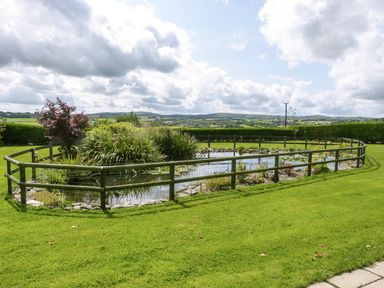 A fenced pond with plants inside and surrounding it in a grassy garden with hills in the background at The Wagon House in Polyphant near Launceston