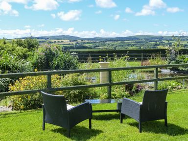 Two outdoor chairs with a glass-top table on grass near a wooden fence and garden at The Wagon House in Polyphant near Launceston