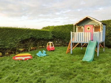 A children's play area with a small playhouse with a slide a red toy car a blue rocking horse and a red sandbox on grass at The Wagon House in Polyphant near Launceston