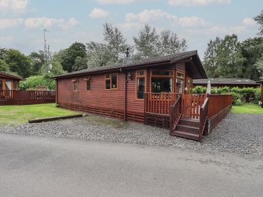 A wooden cabin with a fenced porch and gravel driveway surrounded by trees at Langdale Lodge 15 White Cross Bay near Troutbeck Bridge
