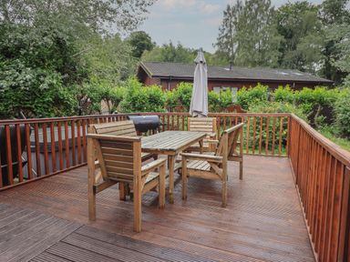 An outdoor wooden deck with a table and four chairs an umbrella and a barbecue grill at Langdale Lodge 15 White Cross Bay near Troutbeck Bridge