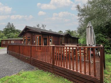 An outdoor wooden deck with patio furniture and a closed umbrella attached to a brown wood cabin at Langdale Lodge 15 White Cross Bay near Troutbeck Bridge