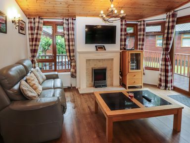 A living room with a gray sofa a coffee table a fireplace a wall mounted tv and windows with plaid curtains at Langdale Lodge 15 White Cross Bay near Troutbeck Bridge