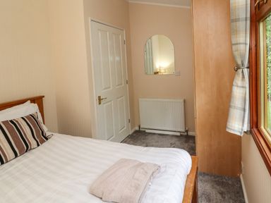 A bedroom with a bed, folded towels on the bed, a door, a wall mirror, a radiator, a wardrobe, and a window with plaid curtains at Langdale Lodge 15 White Cross Bay near Troutbeck Bridge