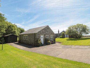 A stone building with a white door and windows next to a wooden shed and a large lawn with trees at Drovers Rest in Otterburn