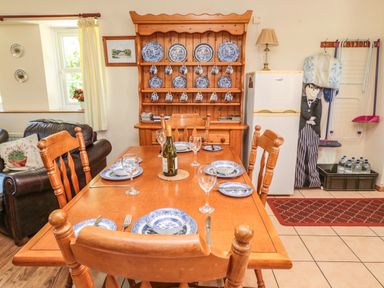 A dining area with a wooden table set with plates, glasses, and a bottle, wooden chairs, a wooden plate rack with blue and white dishware, a refrigerator, and a small living area in the background at Drovers Rest in Otterburn