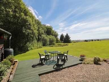 A wooden deck with a glass table and four chairs beside a gravel area and a large grassy field at Drovers Rest Otterburn