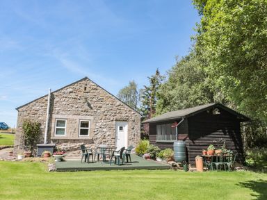 A stone house with a white door and windows next to a wooden shed with outdoor furniture and plants on a grassy lawn at Drovers Rest in Otterburn