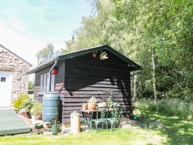 A small black wooden shed with a rain barrel and outdoor table with chairs and potted plants in a garden at Drovers Rest in Otterburn