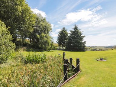 A grassy garden area with trees a small pond and a rope fence at Drovers Rest in Otterburn
