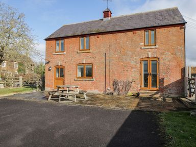 A brick house with wooden windows and doors and a wooden picnic table outside on a paved area at Stables Cottage in Bowerhill near Melksham
