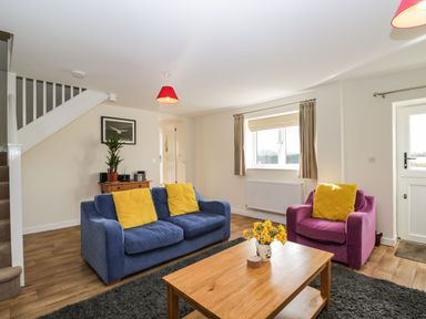 A living room with blue and purple sofas with yellow cushions a wooden coffee table with flowers and a staircase at Stables Cottage in Bowerhill near Melksham
