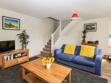 A living room with a blue sofa and yellow cushions a wooden coffee table with flowers television on wooden stand and stairs at Stables Cottage in Bowerhill near Melksham