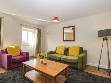 A living room with a green sofa and a purple armchair with yellow cushions a wooden coffee table with flowers and a floor lamp at Stables Cottage in Bowerhill near Melksham
