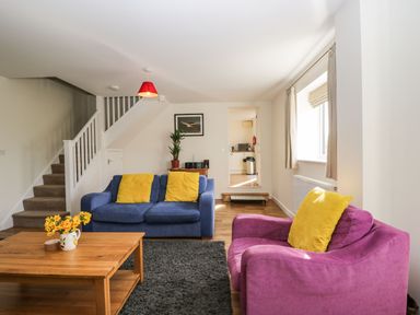A living room with a blue sofa and a purple armchair with yellow cushions a wooden coffee table with flowers and a staircase in the background at Stables Cottage in Bowerhill near Melksham