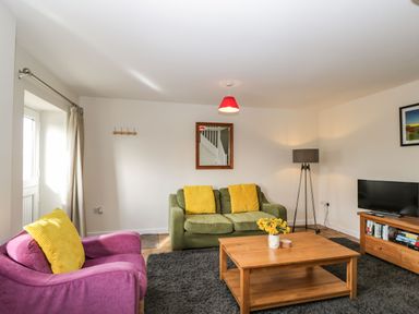 A living room with a green sofa with yellow cushions a purple armchair with a yellow cushion a wooden coffee table with flowers a floor lamp a television on a wooden stand and a gray rug at Stables Cottage in Bowerhill near Melksham