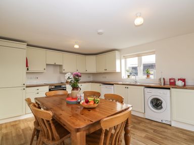 A kitchen with wooden table and chairs a washing machine a dishwasher and cabinets with a window above the sink at Stables Cottage in Bowerhill near Melksham