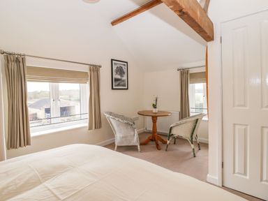 A bedroom with a bed two wicker chairs a round wooden table and windows with beige curtains at Stables Cottage in Bowerhill near Melksham