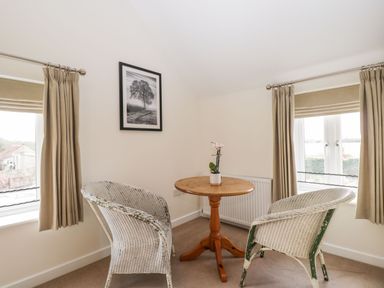 A small dining area with a round wooden table between two white wicker chairs under two windows with beige curtains at Stables Cottage in Bowerhill near Melksham