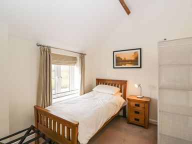 A bedroom with a single wooden bed beside a wooden nightstand with a lamp and a window with beige curtains at Stables Cottage in Bowerhill near Melksham