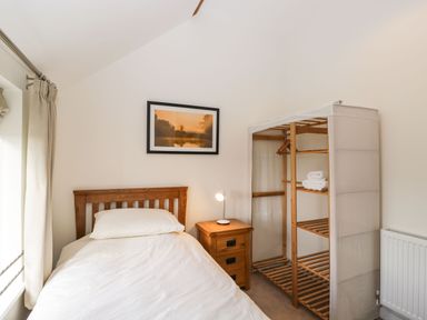 A bedroom with a single bed wooden headboard wooden nightstand with lamp open wardrobe with shelves and towels a radiator on the wall and a framed picture above the bed at Stables Cottage in Bowerhill near Melksham