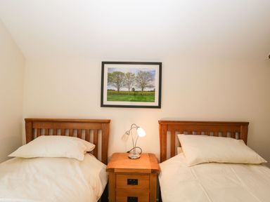 A bedroom with two single beds separated by a wooden nightstand with a lamp and a framed picture of trees on the wall at Stables Cottage in Bowerhill near Melksham