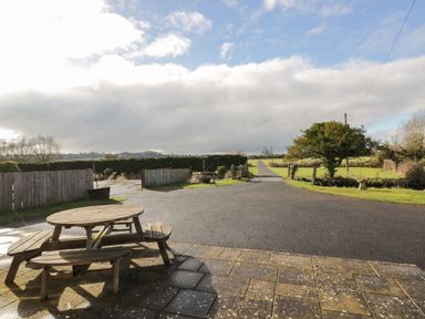 A wooden picnic table on a paved patio overlooking a driveway with fields and hedges at Stables Cottage in Bowerhill near Melksham