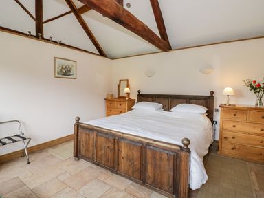 A bedroom with a wooden bed and white bedding two wooden dressers with lamps and flowers exposed wooden ceiling beams at Beesoni Lodge in Castlemorton near Malvern