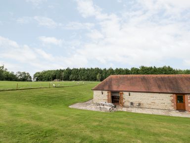 A stone building with tiled roof next to a patio table and chairs on a lawn with sheep grazing in a fenced field and trees in the background at Beesoni Lodge in Castlemorton near Malvern