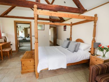 A bedroom with a wooden four-poster bed a bedside table with a lamp and flowers a wooden desk and chair and exposed ceiling beams at Beesoni Lodge in Castlemorton near Malvern