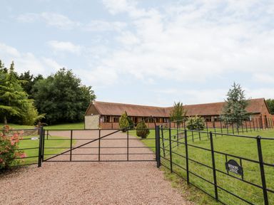 A gravel driveway with a black metal gate leading to a single-story brick and stone building with a pitched roof surrounded by grass and trees at Beesoni Lodge in Castlemorton near Malvern