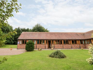 A single-story brick building with multiple windows and a tiled roof surrounded by grass and trees at Beesoni Lodge in Castlemorton near Malvern