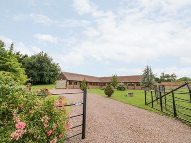 A gravel driveway with a black metal gate leading to a single-story building with a tiled roof and brick walls at Beesoni Lodge in Castlemorton near Malvern