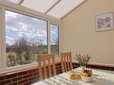 A dining area with wooden chairs a table with fruit and flowers near a large window at 1 Cowdea Farm in Bettiscombe near Beaminster and Bridport