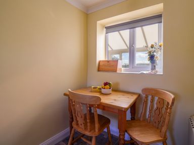 A small dining area with a wooden table and two chairs next to a window with flowers and a bread box on the windowsill at 1 Cowdea Farm in Bettiscombe near Beaminster and Bridport