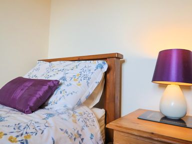 A bedroom with a wooden headboard bed with floral bedding and a purple pillow and a wooden bedside table with a white lamp with a purple shade at 1 Cowdea Farm in Bettiscombe near Beaminster and Bridport