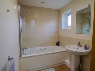 A bathroom with a bathtub and shower, a pedestal sink, a mirror, a window, and a towel hanging on the door at 1 Cowdea Farm in Bettiscombe near Beaminster and Bridport