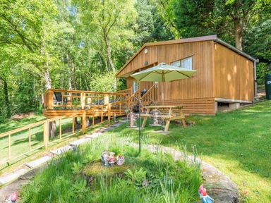 A wooden cabin with a deck and picnic table with umbrella surrounded by trees at Bluebell Cottage in Maenan near Llanrwst