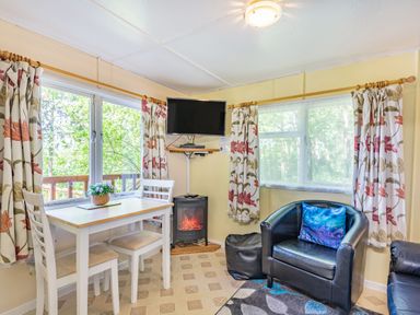 A small dining table with two chairs beside windows with floral curtains a wall-mounted television an electric fireplace a black armchair with a blue cushion and a window at Bluebell Cottage in Maenan near Llanrwst