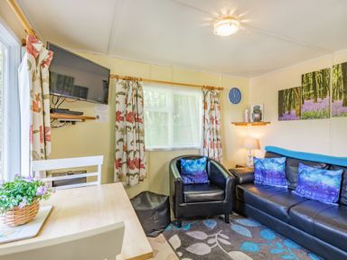 A living room with a black leather sofa and chair blue pillows a wall clock TV on the wall and a wooden table with chairs at Bluebell Cottage in Maenan near Llanrwst