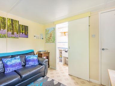A living room with a black leather sofa with blue cushions a small wooden side table artwork on the wall and a sliding door leading to a kitchen at Bluebell Cottage in Maenan near Llanrwst