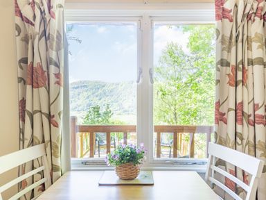 A dining area with a wooden table and white chairs by a window with floral curtains and a view of trees and hills at Bluebell Cottage in Maenan near Llanrwst