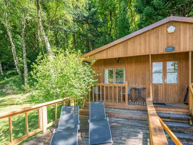 Outdoor deck with two lounge chairs and a wooden cottage surrounded by trees at Bluebell Cottage in Maenan near Llanrwst