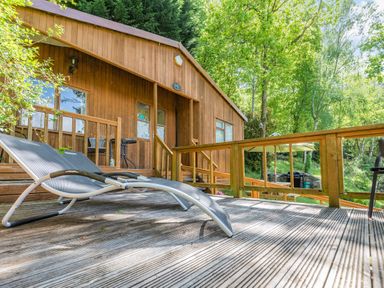 A wooden deck with lounge chairs and a table outside a wooden cabin surrounded by trees at Bluebell Cottage in Maenan near Llanrwst