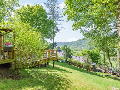 A grassy garden with trees and a wooden deck overlooking hills at Bluebell Cottage Maenan near Llanrwst