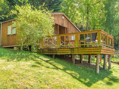 A wooden cabin with a raised deck and outdoor chairs surrounded by trees and grass at Bluebell Cottage in Maenan near Llanrwst
