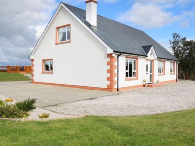 A white house with orange trim and a gray roof on a gravel driveway with grass and shrubs at Sallywood House in Carn near Ballybofey County Donegal