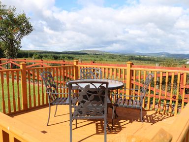 An outdoor wooden deck with a round glass table and four metal chairs overlooking fields and hills at Sallywood House in Carn near Ballybofey County Donegal